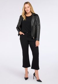 Donna con capelli biondi che indossa una giacca di pelle nera, una blusa nera a pois, pantaloni neri a vita alta e scarpe col tacco a punta nere, in piedi con atteggiamento sicuro.