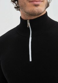 Pull en maille noir avec un col montant et une fermeture éclair argentée. Présente un détail de fermeture éclair blanc contrastant et un matériau texturé.