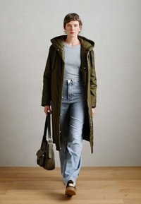 Lange olijfgroene parka met een capuchon en rits, gedragen over een lichtgrijze top en hoge taille blauwe jeans. Bruine schoenen en een schoudertas inbegrepen.