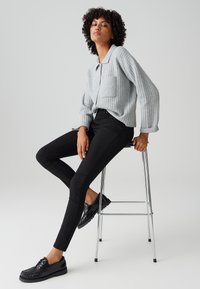 Femme aux cheveux bouclés assise sur un tabouret en métal, portant un pull gris côtelé, un pantalon skinny noir et des mocassins noirs, sur un fond gris uni.