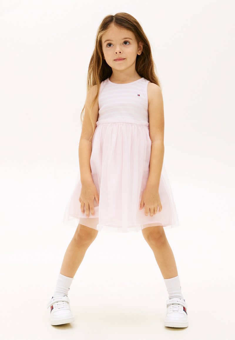 Mladé dievča s dlhými vlasmi, oblečené v ružových šatách bez rukávov a bielych teniskách, stojace s nohami od seba na jednoduchom pozadí.