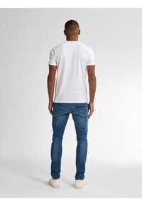 Vit t-shirt med korta ärmar, blå denimjeans med sydda bakfickor och vita sneakers, sedd bakifrån mot en ljus bakgrund.