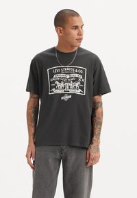 T-shirt en coton noir avec un imprimé graphique blanc comprenant un logo et un artwork de style vintage ; coupe décontractée, col rond, manches courtes.