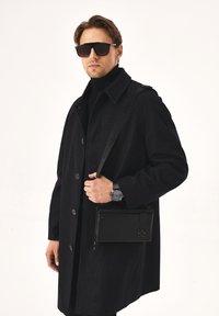 Fekete kabát texturált felülettel, turtleneck fölött viselve. Fekete táska logóval a kezében, sötét napszemüveggel és órával párosítva.