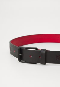 Ceinture en cuir noir avec intérieur rouge, dotée d'une boucle en métal noir et d'un logo en relief. Comprend plusieurs trous pour un ajustement.