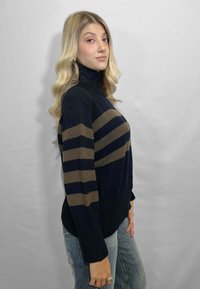 Giovane donna con lunghi capelli biondi, che indossa un maglione a collo alto a righe blu navy e marrone e jeans blu chiaro, in piedi di profilo contro uno sfondo grigio.