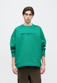 Πράσινο φούτερ με στρογγυλή λαιμόκοψη, άνετη εφαρμογή και ριμπ μανσέτες. Διαθέτει το κείμενο "JUST DO IT." και το λογότυπο της Nike σε ναυτικό μπλε χρώμα.