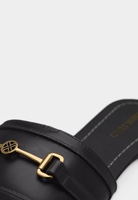 Chaussure en cuir noir à enfiler avec un détail en métal doré et des coutures blanches sur le bord de la semelle, présentée en gros plan sur un fond blanc.