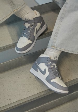 Person iført hvide og grå Nike Air Jordan sneakers og lyse jeans, siddende på grå trapper.