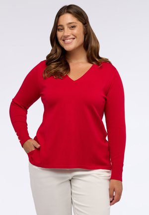 Donna sorridente che indossa un maglione rosso a V con polsini bottonati e pantaloni bianchi, che posa con una mano in tasca contro uno sfondo bianco.