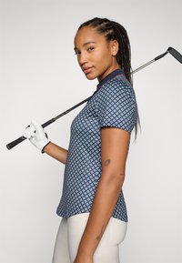 Zilās golf polo krekls ar ģeometrisku rakstu, īsām piedurknēm un apkakli, apvienots ar gaišām biksēm. Golfu klubs turēts rokā.