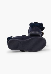 Sandalias de terciopelo azul marino con suela de goma texturizada, que presentan un gran lazo de terciopelo en la parte trasera y una correa con cierre de hebilla.