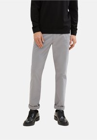 Graue Baumwoll-Chinos mit geradem Schnitt, umgeschlagenen Säumen und Fronttaschen, kombiniert mit schwarzen Schnürschuhen und einem schwarzen Pullover.