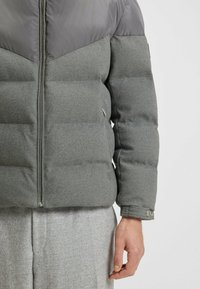 Grå puffjacka med en stilren kviltad design, en slät grå övre del, textil nedre del i grått, och dragkedja.