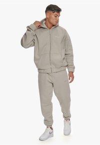 Beige zip-up hoodie with a large hood and front pockets, matched with tapered sweatpants; both in a soft, textured fabric.