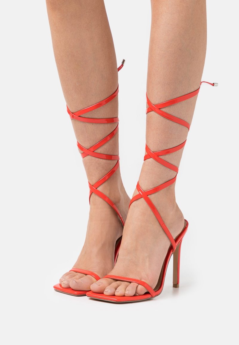 Pieds chaussés de sandales rouges à talons hauts avec lanières enserrant la cheville et bouts carrés ouverts sur fond blanc.