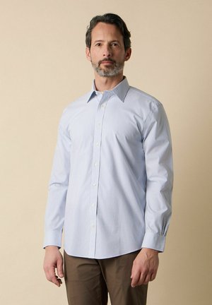 Mann mit salt-and-pepper Bart trägt ein hellblaues, fein gestreiftes Hemd mit Knöpfen und braune Hose und steht vor einem beigen Hintergrund.