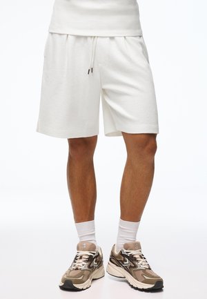 Férfi lábak fehér térdig érő rövidnadrágban, fehér sportzokniban, bézs mintás futócipőben, fehér háttér előtt.