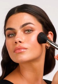 Un pincel de maquillaje con cerdas negras aplica rubor en una mejilla. La persona tiene piel suave, labios llenos y cabello castaño medio.