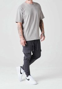 Šedé tričko s krátkým rukávem v kombinaci s černými cargo kalhotami. Oblečení zahrnuje bílé tenisky s černými detaily a na pažích jsou viditelné tetování.