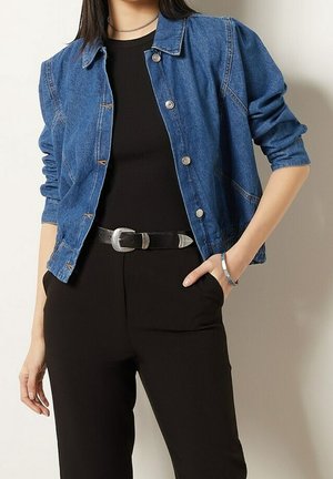 Osoba nosząca niebieską dżinsową kurtkę na czarnym topie, czarne spodnie z paskiem z srebrną, westernową klamrą oraz srebrną bransoletkę na prawym nadgarstku.