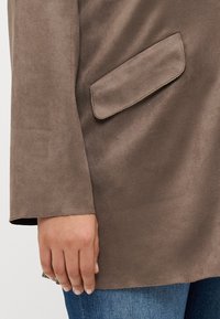 Brown suede fabric with a smooth texture, featuring a side pocket and a straight cut sleeve. The hand shows a natural nail finish.
