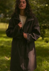 Cappotto impermeabile scuro con vita regolabile tramite cordino, ampie tasche e colletto alto, posizionato su uno sfondo verde e naturale.
