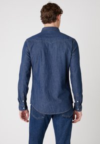 Donkerdenim shirt met lange mouwen en een puntige kraag. Heeft stikseldetails en knoopmanchetten. Achterzoom is licht gebogen.