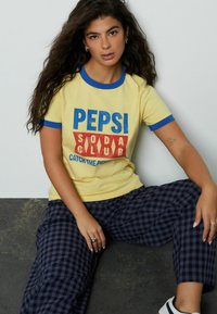 Geel T-shirt met een blauwe runderkraag, met een rood-blauwe "PEPSI SODA CLUB" afbeelding. Gecombineerd met marineblauwe geruite broek. Textuur is glad.