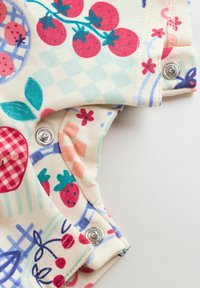 Bombažna tkanina za otroško oblačilo v svetlo kremni barvi z rdečimi paradižniki, jagodami in modrimi detajli. Na strani so pritiskači za zapenjanje.