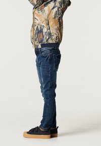 Barn som bär en zip-up jacka med blommönster, blå jeans med slim fit och svarta skor med gum-sula.