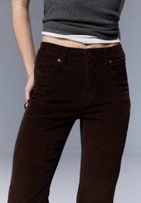 Pantalon en velours côtelé marron avec une surface texturée et côtelée. Dispose d'une taille ajustée et de poches avant, fermées par des boutons en métal.