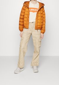 Doudoune orange en tissu texturé ; t-shirt crème avec logo Levi's rouge ; pantalon cargo beige avec motif marbré ; baskets gris clair.