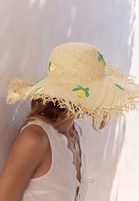 Femme portant un grand chapeau de paille orné d'ananas brodés et un débardeur blanc contre un mur clair.
