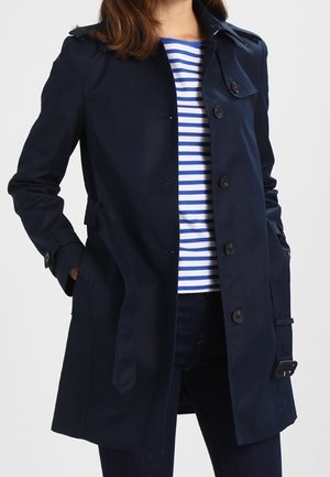 Vrouw draagt een marineblauwe trenchcoat over een wit-blauw gestreept shirt, met haar handen in de zakken van de jas.