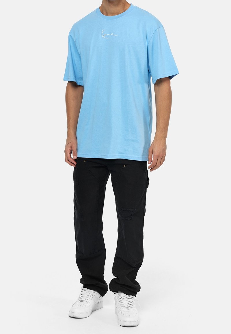 Ljusblå bomulls t-shirt med en liten vit logotyp på bröstet, ihop med svarta cargo-byxor och vita sneakers.