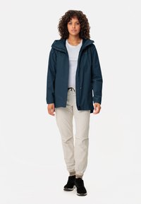 Marineblaue wasserdichte Jacke mit Kapuze, Frontreißverschluss und verstellbaren Kordeln, getragen über einem hellgrauen Shirt und hellbeigen Hosen.