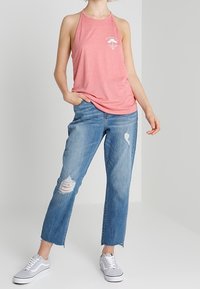 Lichtroze haltertop met een witte grafische print, gedragen met vervaagde blauwe jeans met slijtage en rauwe randen, gecombineerd met grijze sneakers.