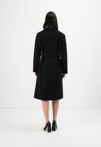 Donna in piedi di spalle, che indossa un cappotto nero lungo fino al ginocchio con cintura e scarpe con tacco alto nere, su uno sfondo bianco semplice.