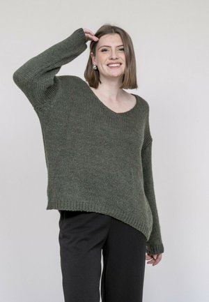 Femme souriante portant un pull ample vert olive et un pantalon noir, levant un bras avec les doigts touchant sa tête sur un fond uni.