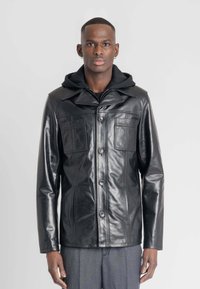 Zwarte leren jas met een kraag, twee borstzakken en een knopenfront. Daaronder is een zwart hoodie zichtbaar.