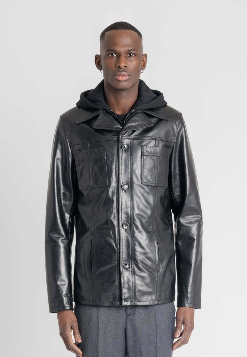 Zwarte leren jas met een kraag, twee borstzakken en een knopenfront. Daaronder is een zwart hoodie zichtbaar.