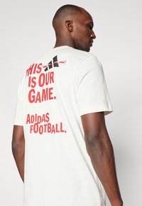 Hvid bomulds T-shirt med iøjnefaldende rødt tekst på ryggen: "DETTE ER VORES SPIL. ADIDAS FODBOLD." med et særpræget logodesign.