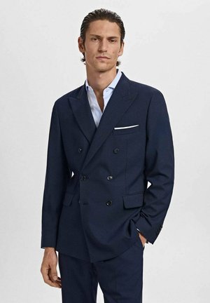 Veston bleu marine à double boutonnage avec des boutons noirs, revers crantés et une pochette blanche. Texture lisse, coupe ajustée.