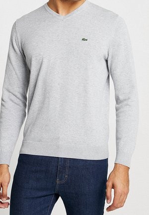 Homme portant un pull gris clair en V et un jean bleu foncé, debout devant un fond clair uni.