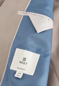 Veste beige slim fit avec doublure bleue montrant l'étiquette de marque cousue "NEXT" et l'étiquette de taille "40R Made in India."