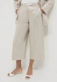 Pantaloni cropped beige lucidi con pieghe, tasche frontali e design a gamba larga, abbinati a tacchi bianchi open-toed.