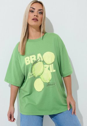 Frau mit langen blonden Haaren, die ein lockeres grünes T-Shirt mit Limettengrafik und dem Schriftzug "Brazil Zesty" sowie blaue Jeans trägt, posiert vor einer weißen Wand.