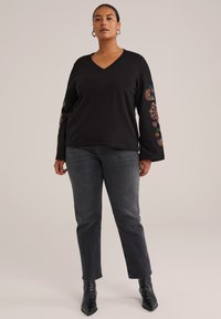 Kvinde iført sort V-hals sweater med blomsterbroderi på ærmerne, mørkegrå jeans og sorte ankelstøvler, der står mod en ensfarvet baggrund.