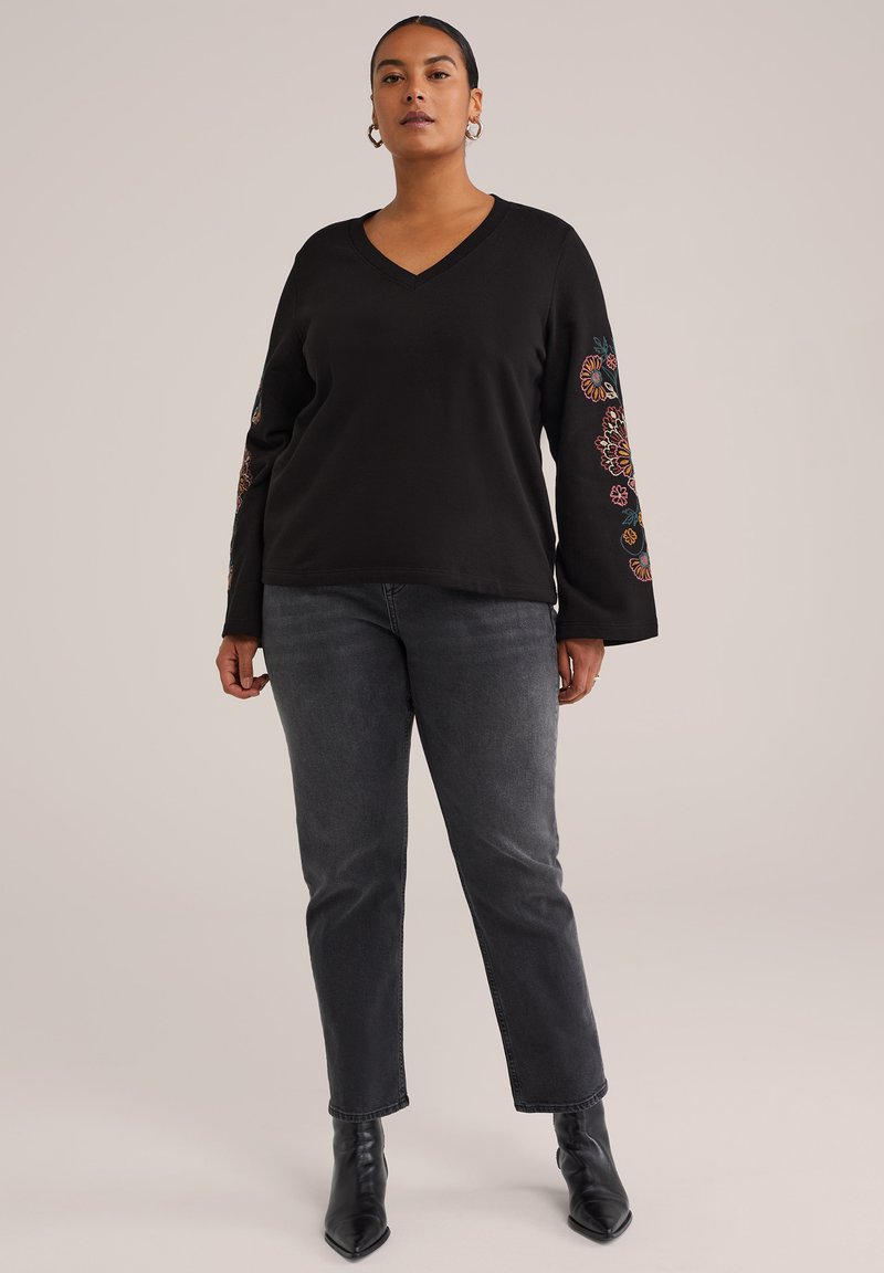 Kvinde iført sort V-hals sweater med blomsterbroderi på ærmerne, mørkegrå jeans og sorte ankelstøvler, der står mod en ensfarvet baggrund.